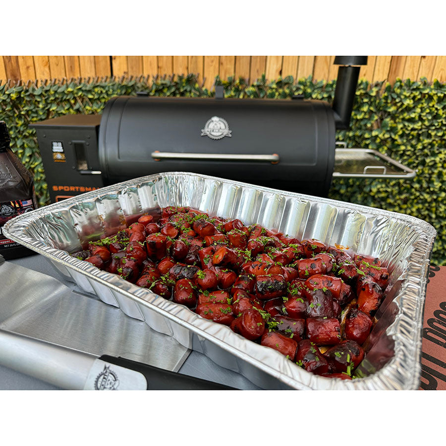 Silver aluminum pan filled with glazed meat chunks, garnished with herbs, near a black grill.