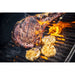 Grilled steak with charred surface, garlic cloves, and flames on a black grill.