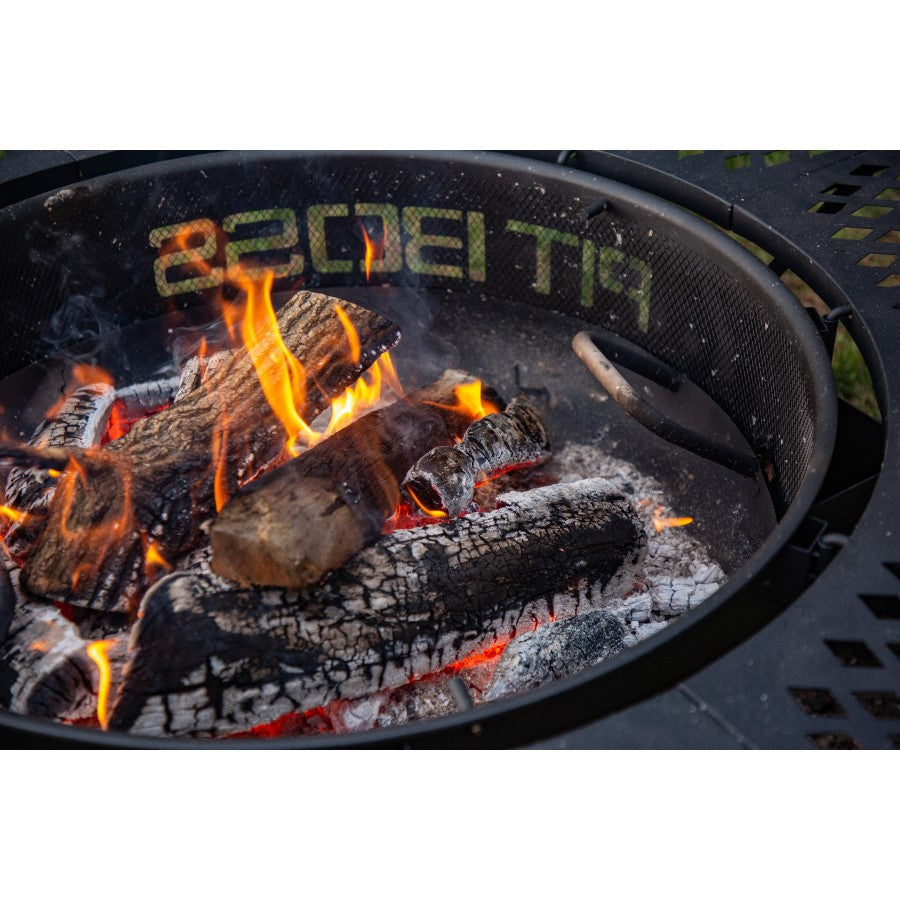 Round metal fire pit with burning logs and flames, mesh sides, and PIT BOSS branding visible on the rim.