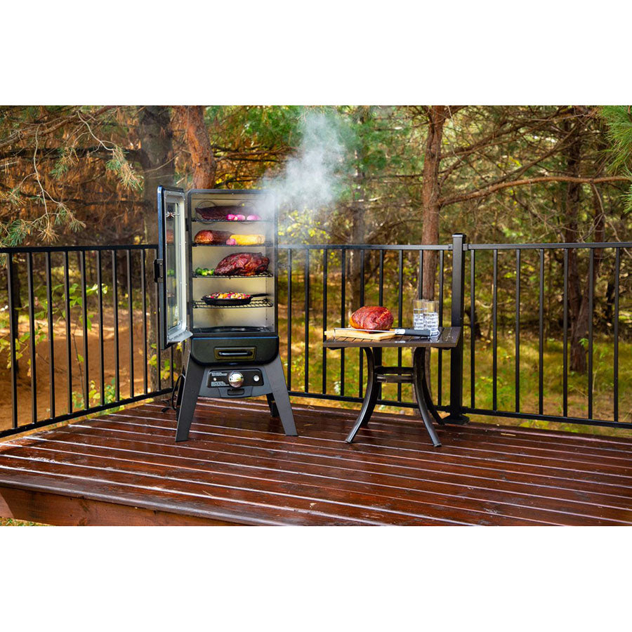 Smoker standing outside on a deck in the trees. Smoker door open. Meats and veggies smoking inside. Mini table displayed next to the smoker with meat and water glasses on it.