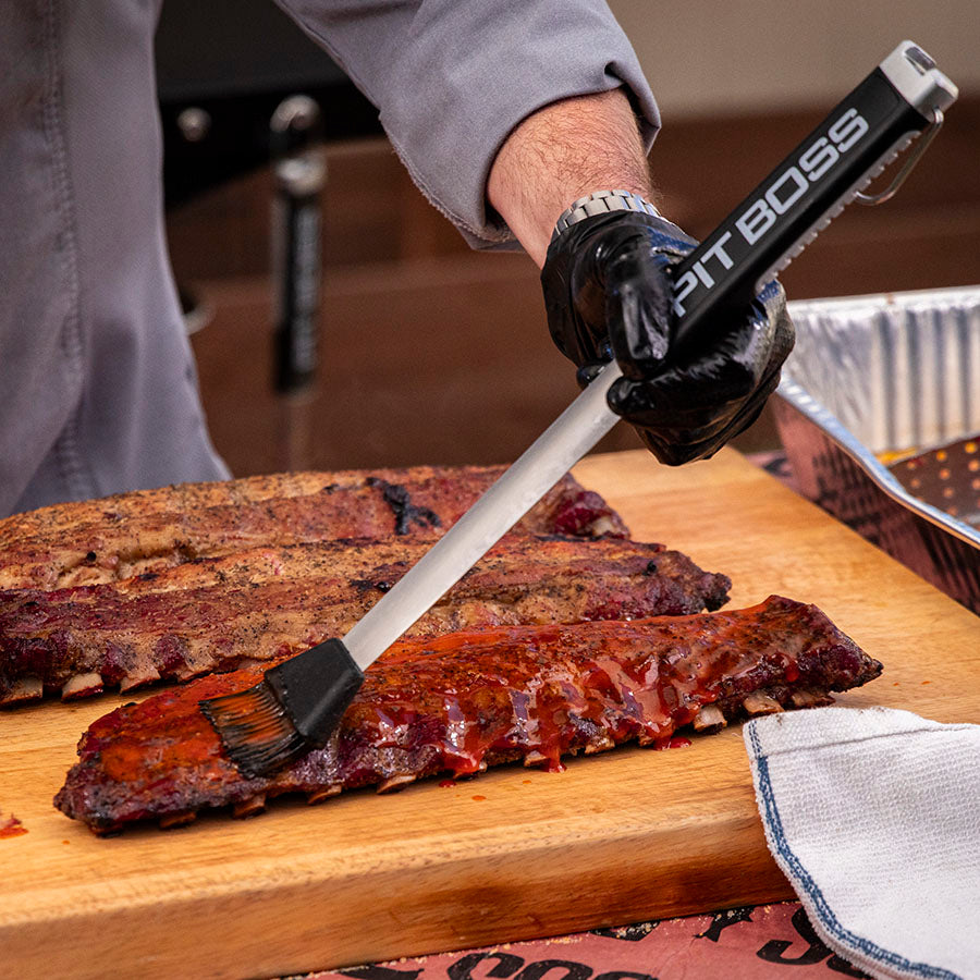 Black Pit Boss BBQ brush applying sauce to ribs on wooden board.