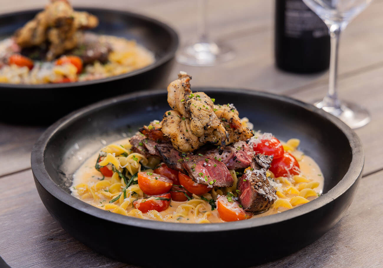 grilled surf and turf with griddled alfredo pasta served on black plate with garnish and cheese.
