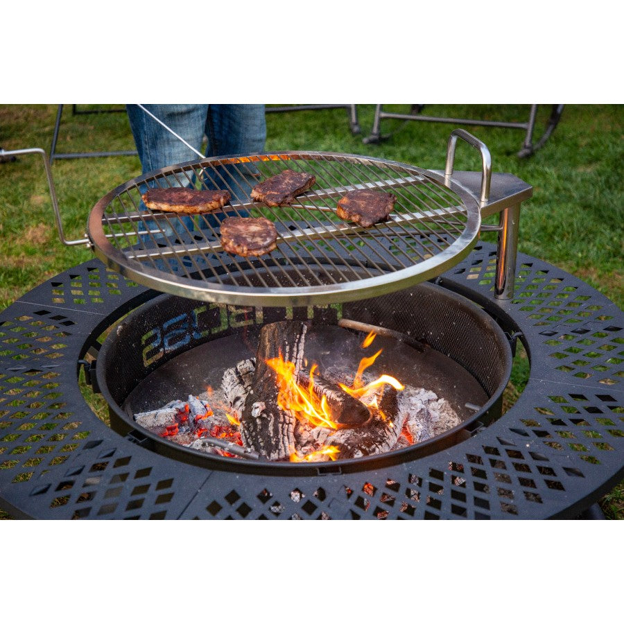Round metal grill grate with burgers cooking over an open fire pit, set on a decorative black metal base outdoors.