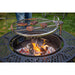 Round metal grill grate with burgers cooking over an open fire pit, set on a decorative black metal base outdoors.