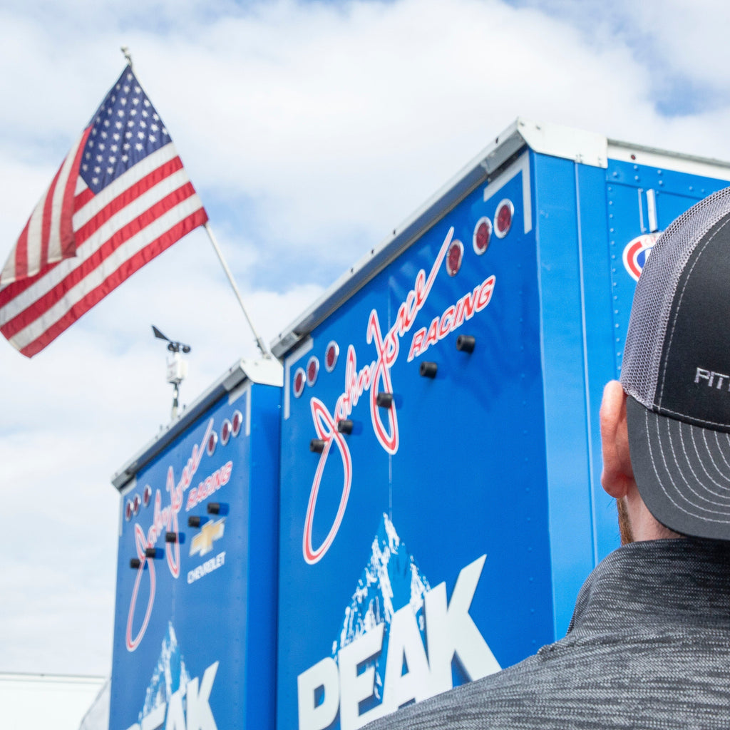 Black PITBOSS trucker hat worn by person near blue racing trailer.