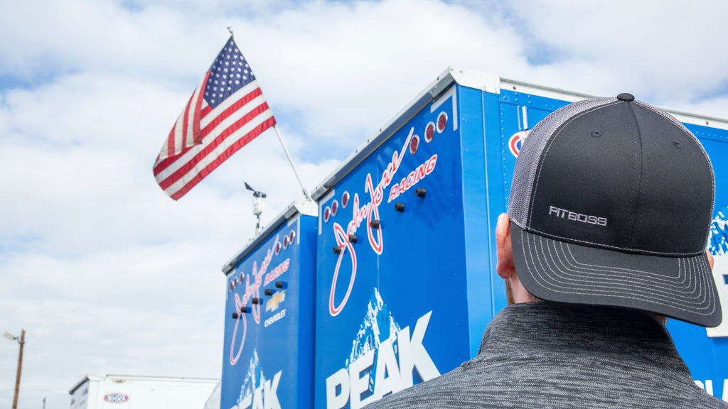 Black PITBOSS trucker hat worn by person near blue racing trailer.