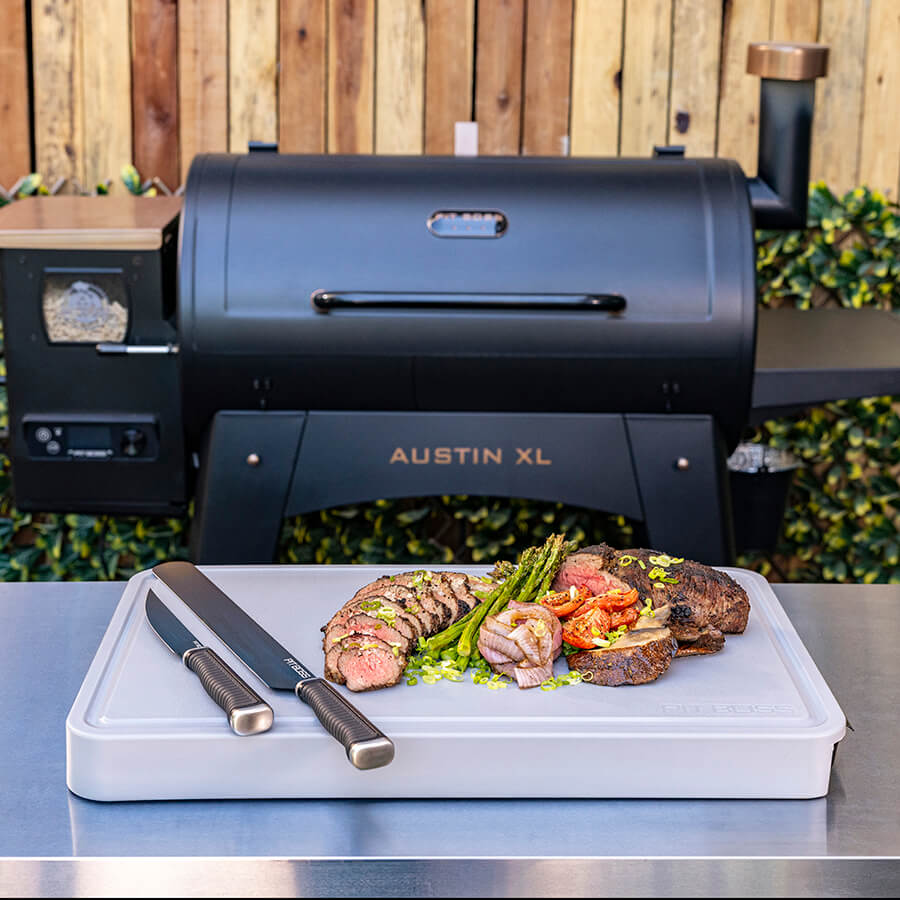 Black Austin XL pellet grill with cooked meats and vegetables on a cutting board.