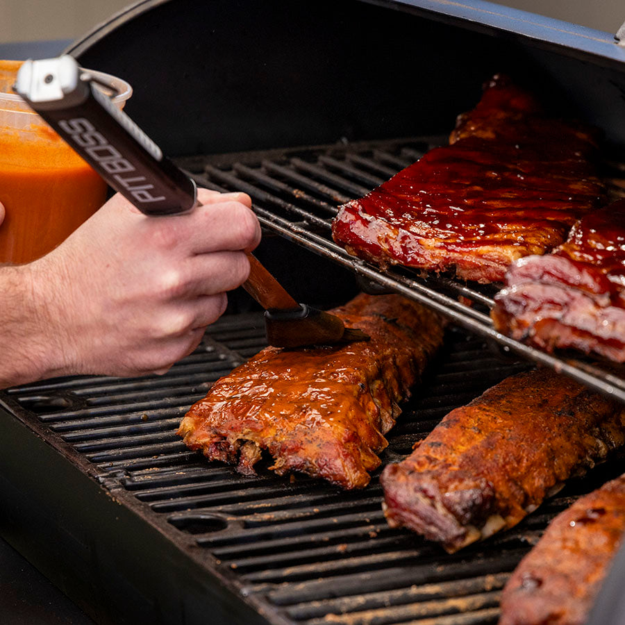 Black grill with ribs being basted with sauce using a Pit Boss brush.
