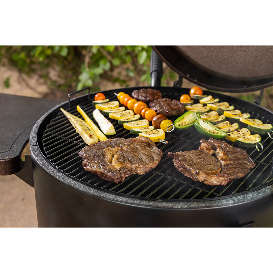 Black round charcoal grill with steaks, vegetables, and burgers cooking.