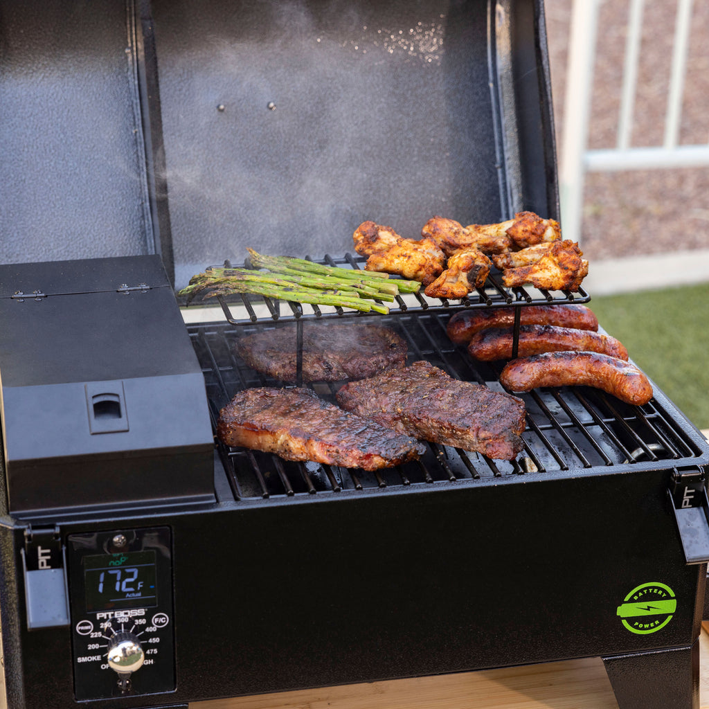 Black electric grill with digital control, grilling steaks, sausages, chicken wings, and asparagus. Open lid, outdoor sett...