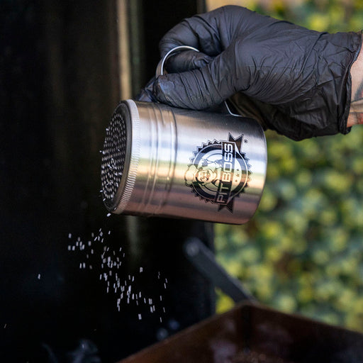 Silver shaker with "Pit Boss" logo, held by gloved hand, dispensing seasoning.