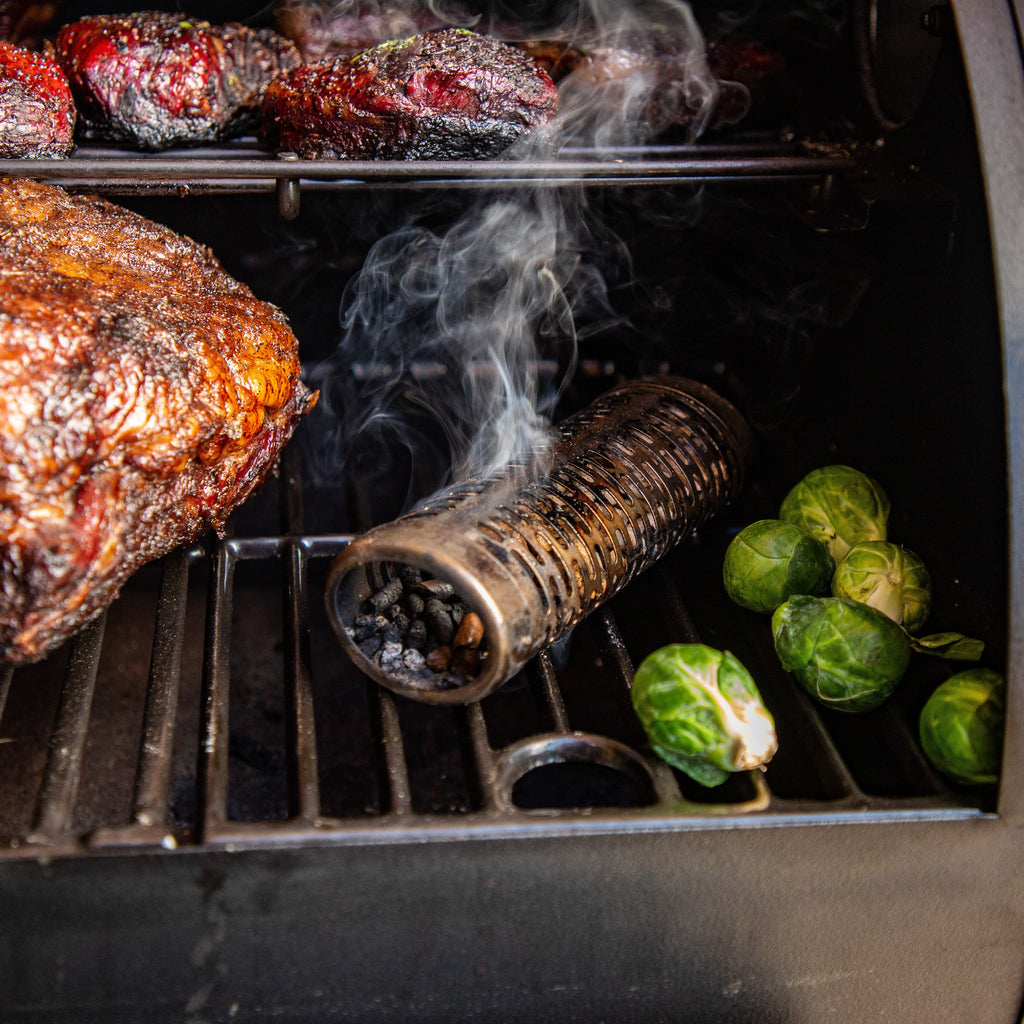 Smoker grill with smoked meat, Brussels sprouts, and a smoking tube releasing smoke on the grill grate.