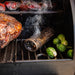 Smoker grill with smoked meat, Brussels sprouts, and a smoking tube releasing smoke on the grill grate.