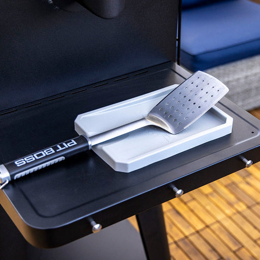 Silver Pit Boss spatula with perforated head, resting on white holder on black grill surface.