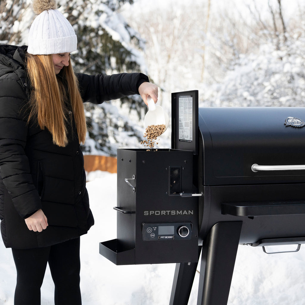 Black Sportsman pellet grill with woman adding pellets in snowy setting.