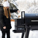 Black Sportsman pellet grill with woman adding pellets in snowy setting.