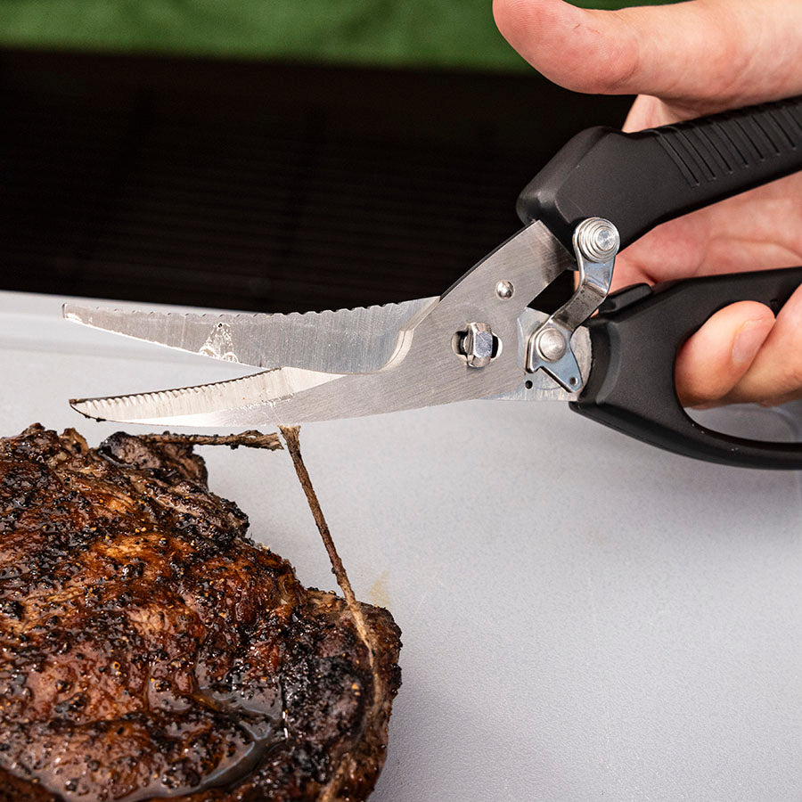 Black-handled kitchen shears cutting into a steak.