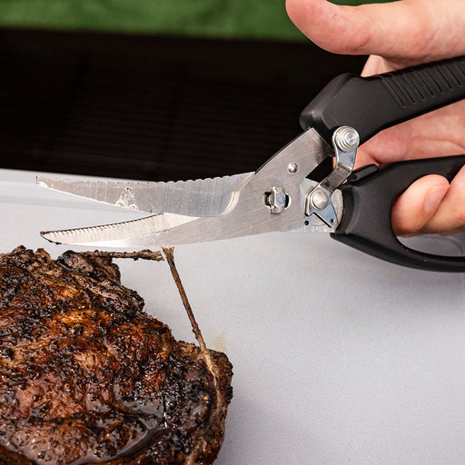 Black-handled kitchen shears cutting into a steak.