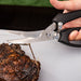 Black-handled kitchen shears cutting into a steak.