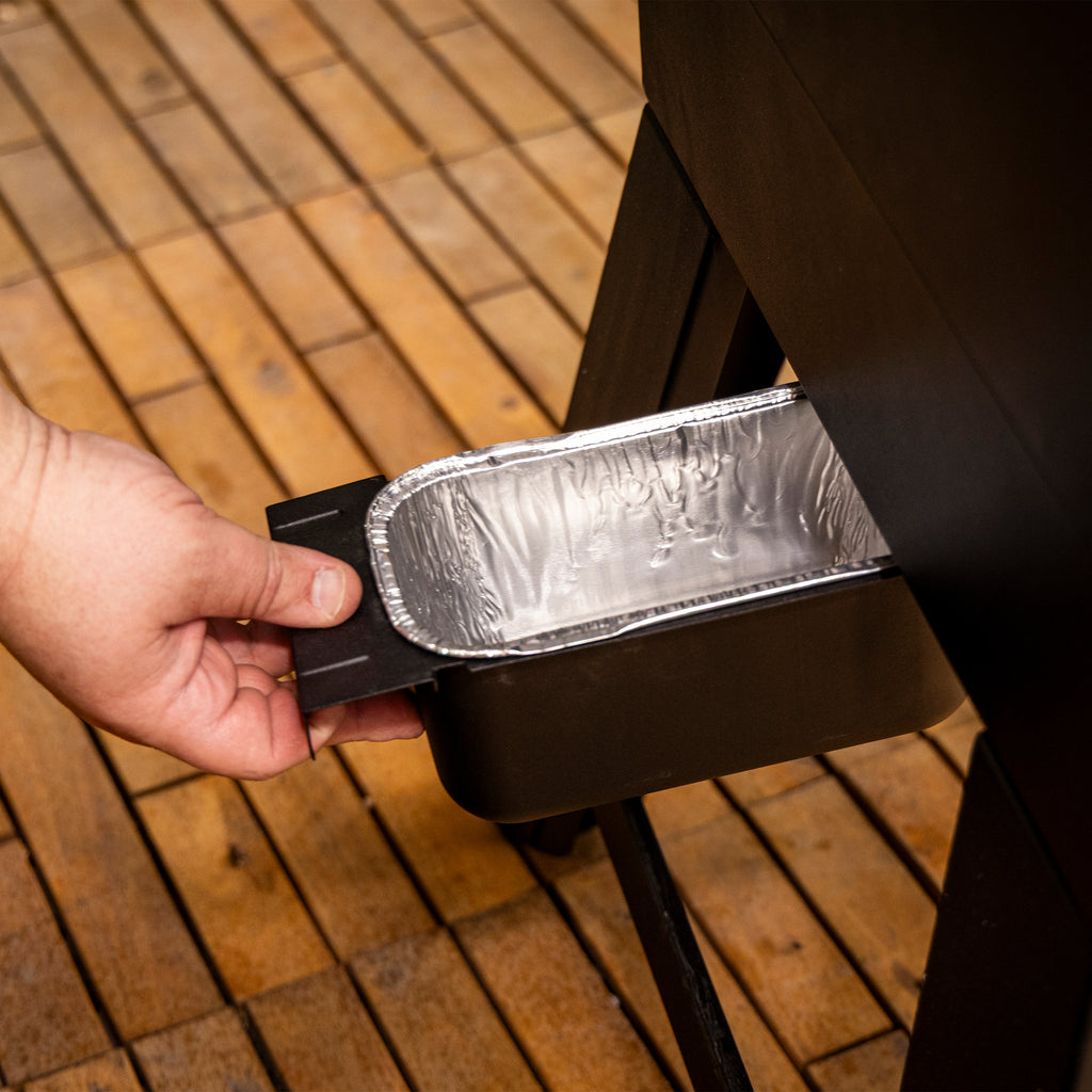 Hand pulling out a rectangular aluminum drip tray from a black grill on a wooden deck. Easy-access grease management.