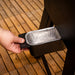 Hand pulling out a rectangular aluminum drip tray from a black grill on a wooden deck. Easy-access grease management.