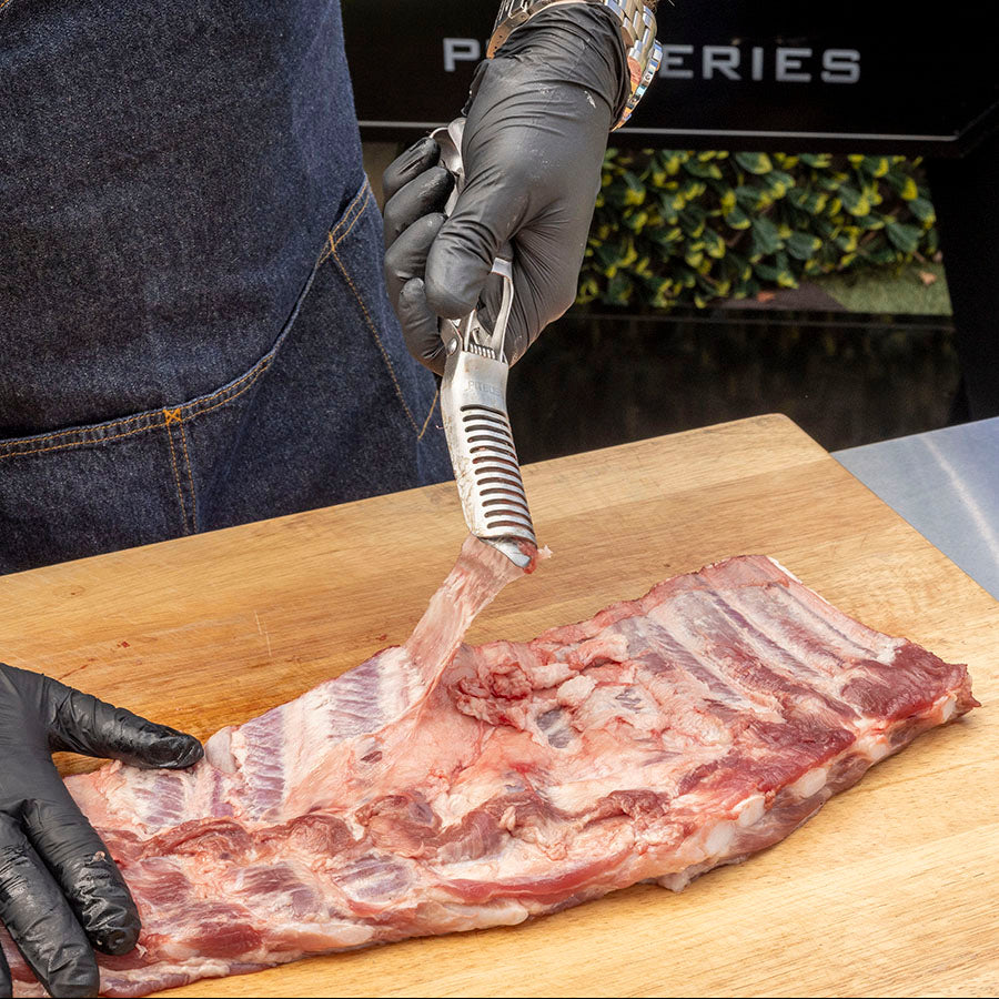Black gloved hands using a silver meat tenderizer on raw ribs on a wooden cutting board.