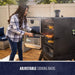 Outdoor smoker with adjustable cooking racks, glass door, and digital controls. Woman grilling meat and corn on patio.