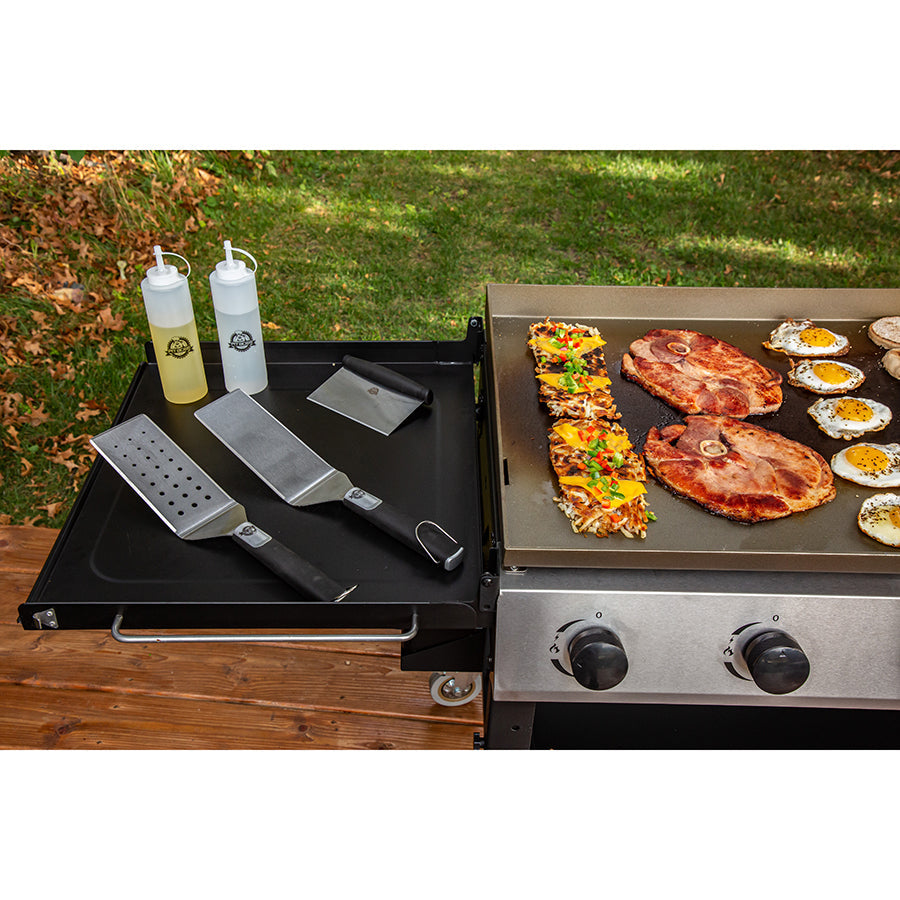 Outdoor cooking station with black griddle, silver tools, and grill cooking steaks, eggs, and veggies.