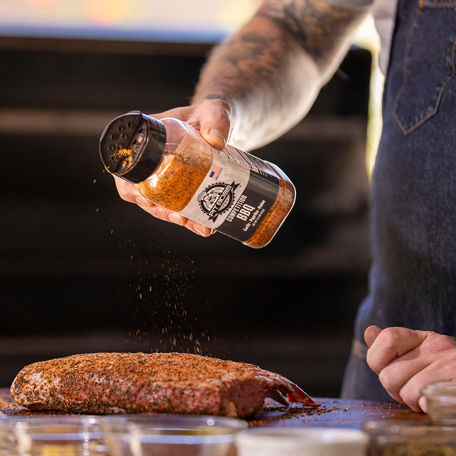 Black "Pit Boss Competition BBQ" spice blend being sprinkled on meat.