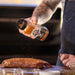 Black "Pit Boss Competition BBQ" spice blend being sprinkled on meat.