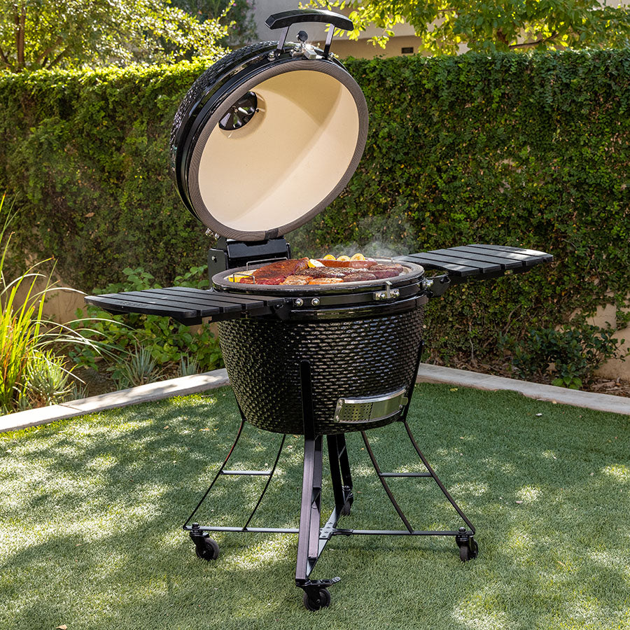 Black kamado-style grill with open lid, cooking food, on wheels, outdoors.