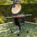 Black kamado-style grill with open lid, cooking food, on wheels, outdoors.