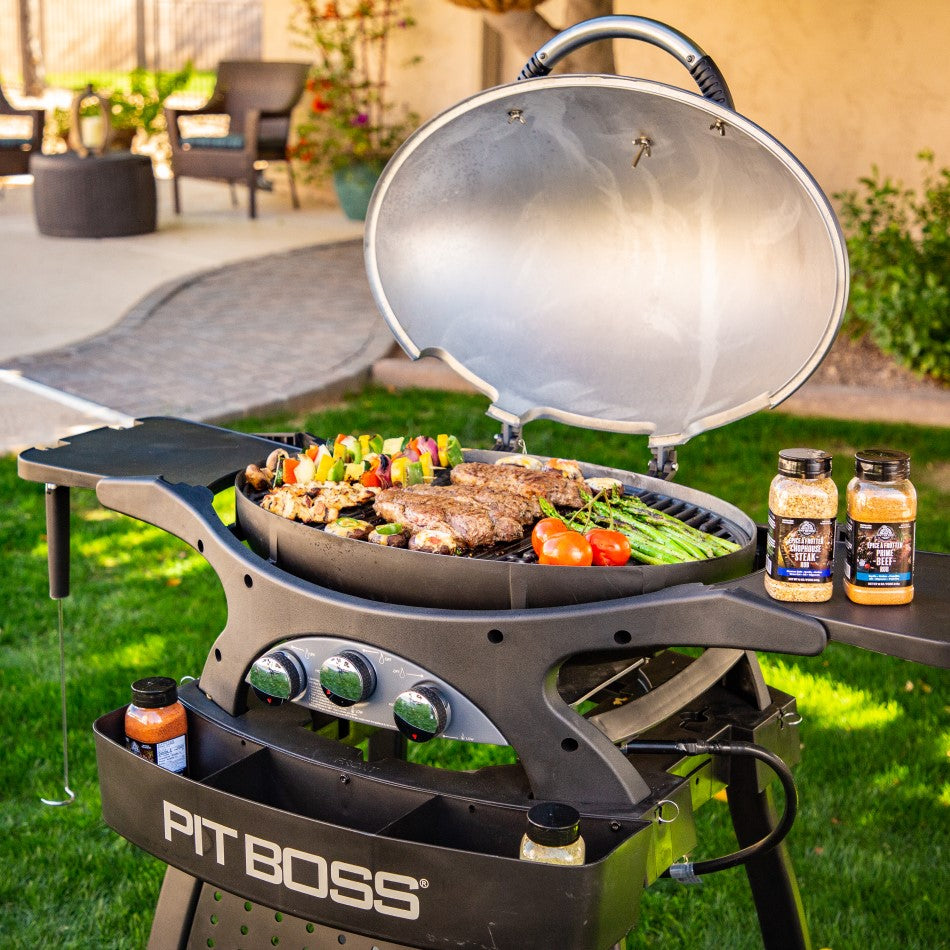 Black Pit Boss portable gas grill with lid open, cooking steaks, vegetables, and seasoning jars nearby.