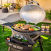 Black Pit Boss portable gas grill with lid open, cooking steaks, vegetables, and seasoning jars nearby.
