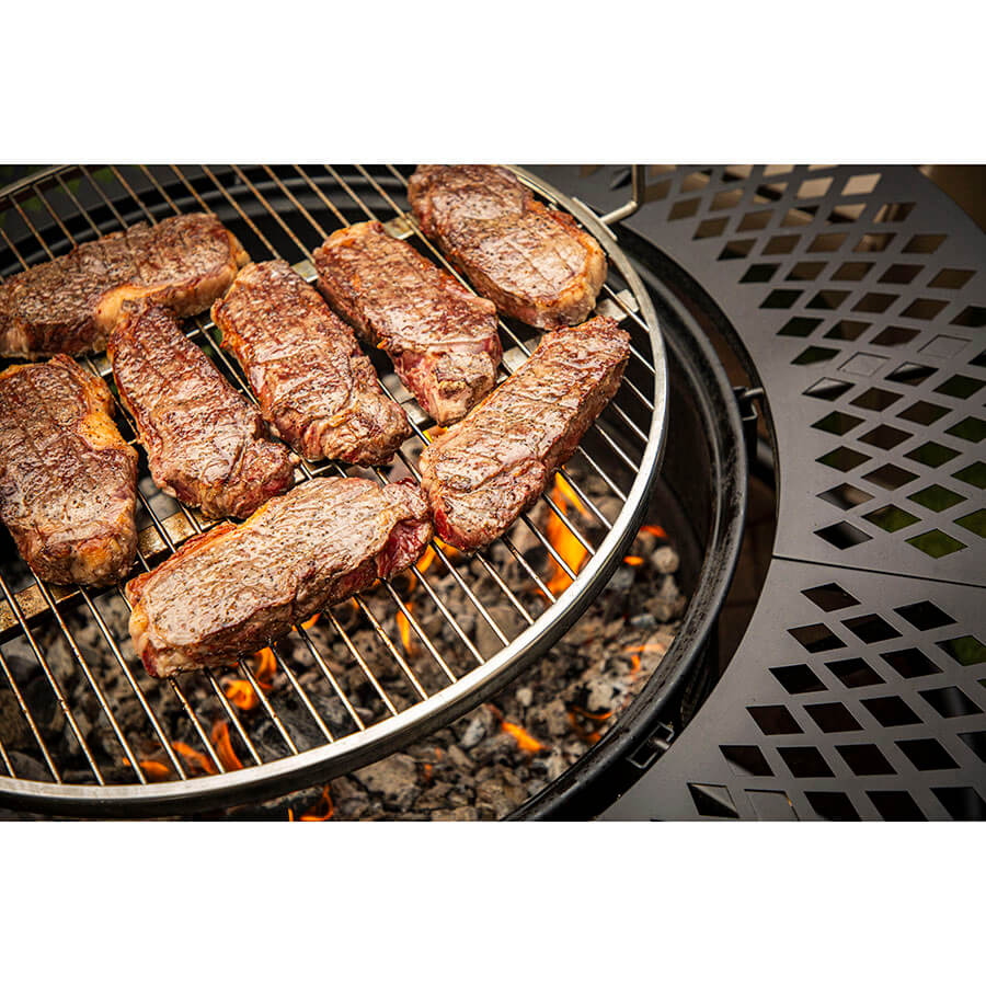 Grill with several steaks cooking over open flames on a round metal grate, surrounded by a patterned metal edge.