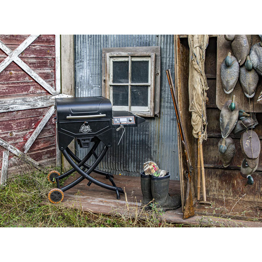 Portable black pellet grill with folding legs and wheels, set on a rustic wooden porch beside boots and hunting gear.