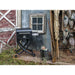 Portable black pellet grill with folding legs and wheels, set on a rustic wooden porch beside boots and hunting gear.